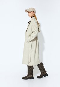 Lang beige frakke med hætte, der har sidelommer. Båret over en brun top. Kombineret med brune knæhøje støvler med spænder. En kasket fuldender looket.