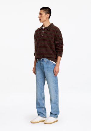 Jeune homme debout en jean bleu clair, polo rayé marron à manches longues et baskets blanches sur fond blanc.