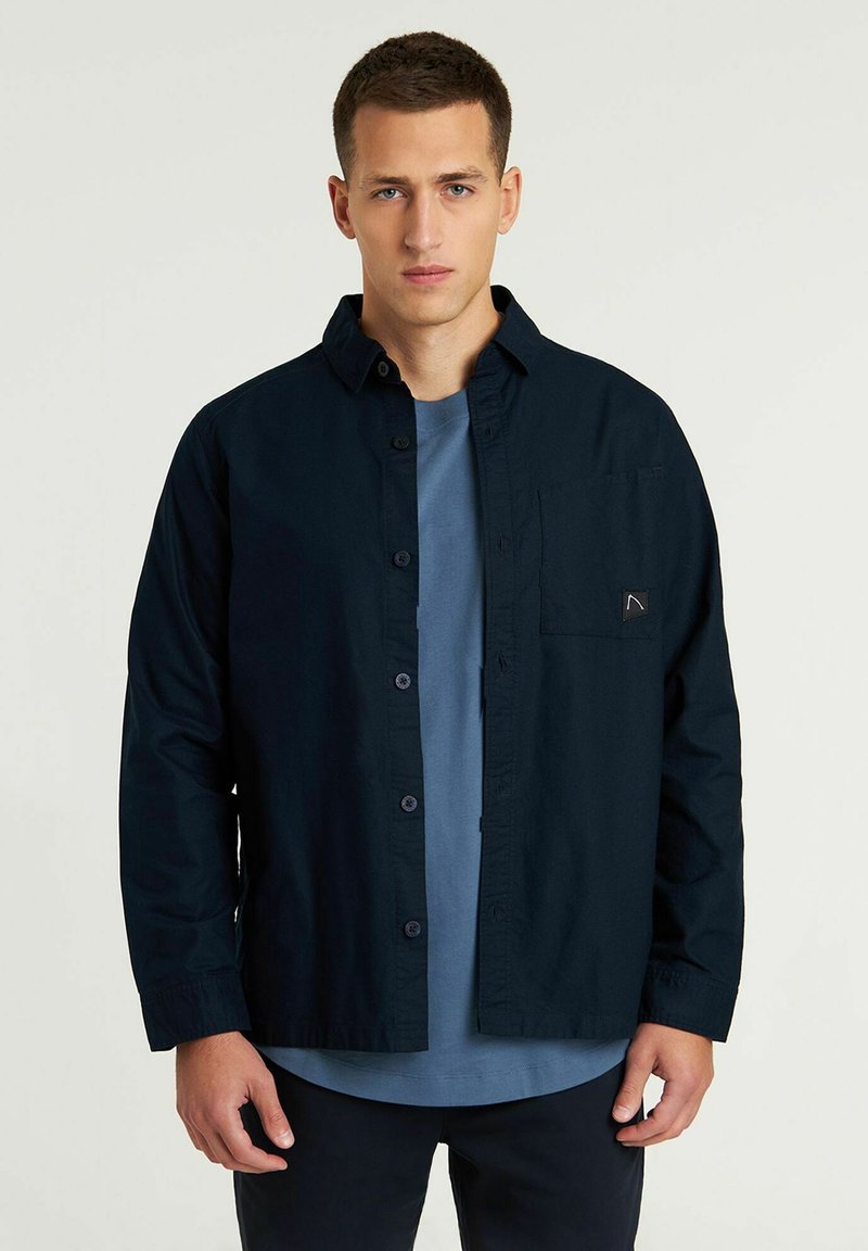 Camisa de botón azul oscuro con un ajuste relajado, que presenta un bolsillo en el pecho y una textura de algodón suave, superpuesta sobre una camiseta azul claro.