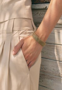 Kuldmetallist ketigaelane käevõru randmel; tekstuuriga, eesmärgiga kujundus. Käsi asetatud beežis pleeditud rõivastes taskusse puidust tausta vastas.