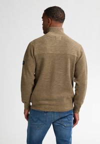 Suéter de punto en beige con cuello alto y tejido texturizado. La vista desde atrás muestra las costuras de los hombros y un parche en la manga izquierda.
