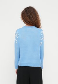 Άτομο με σγουρά μαλλιά που φοράει ανοιχτό μπλε πουλόβερ με λευκά λουλουδένια σχέδια στα μανίκια και μαύρο παντελόνι, με το πρόσωπο στραμμένο προς ένα λευκό φόντο.