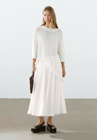 Maglione bianco di maglia oversize abbinato a una lunga gonna bianca fluente e sandali con piattaforma neri. Accessorio: borsa intrecciata marrone.