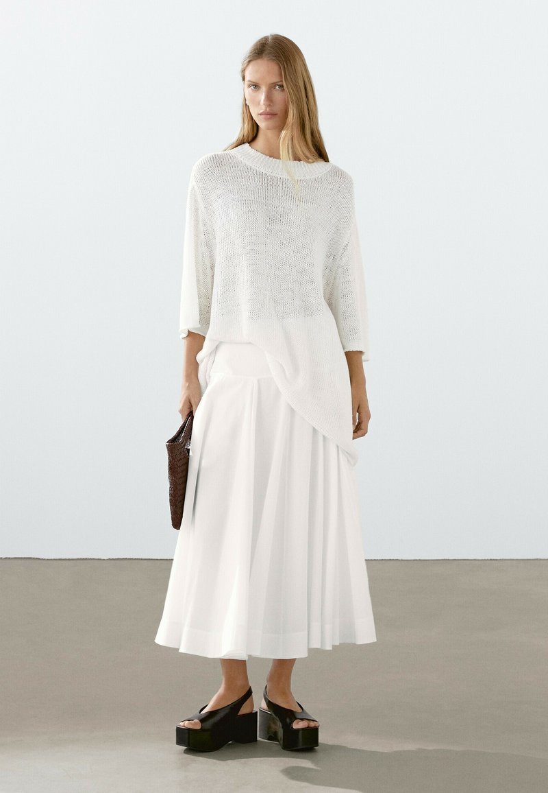 Maglione bianco di maglia oversize abbinato a una lunga gonna bianca fluente e sandali con piattaforma neri. Accessorio: borsa intrecciata marrone.