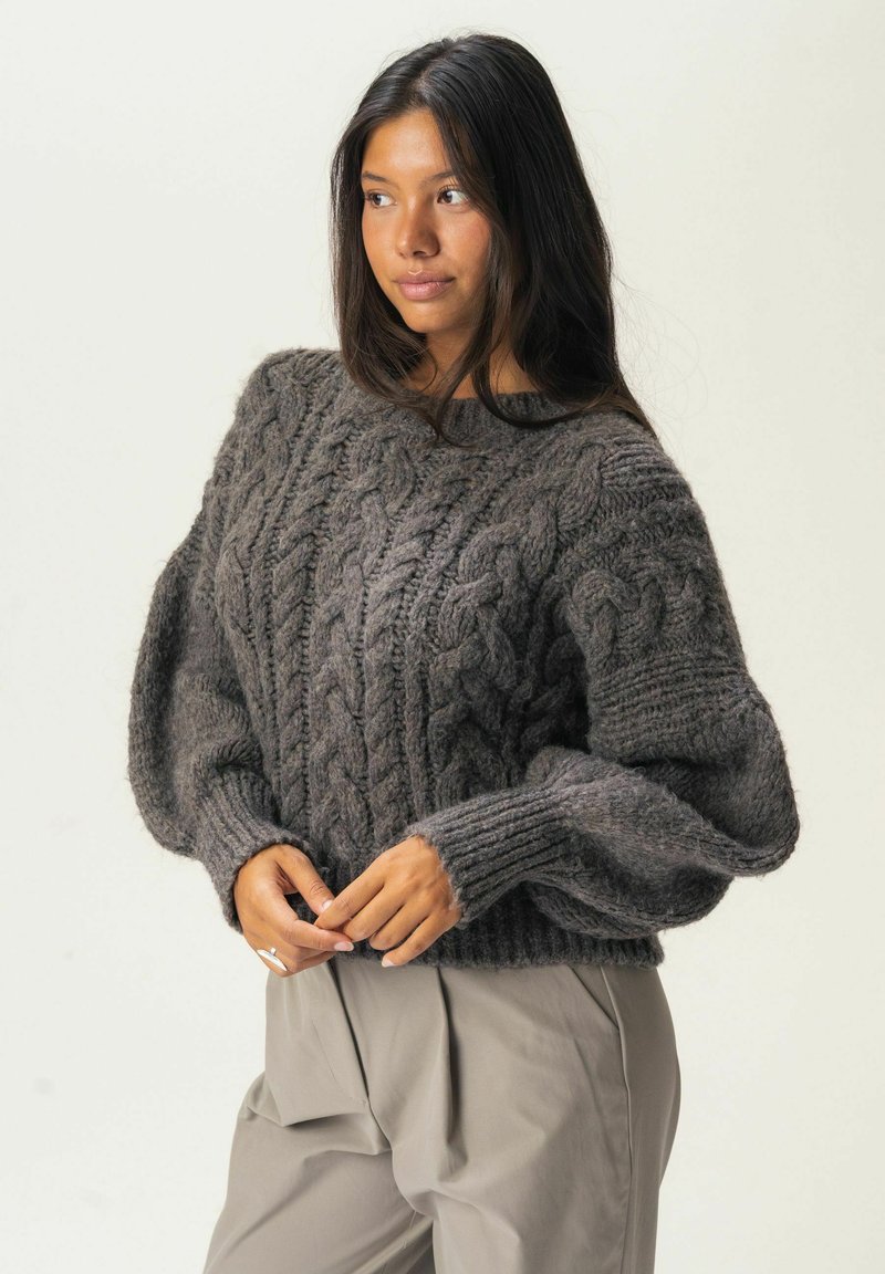 Pull en maille torsadée gris avec des manches larges et des poignets côtelés, fait en fil épais, porté sur un pantalon beige plissé.