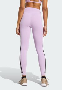 Leggings lilas clairs à taille haute, avec des rayures noires sur les côtés et une texture lisse et extensible. Portés avec des chaussures de sport beiges.