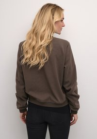 Sweatshirt brun à col rond avec des manches longues, poignets côtelés et une coupe décontractée. Tissu texturé avec une finition lisse et sans motifs ni accessoires visibles.
