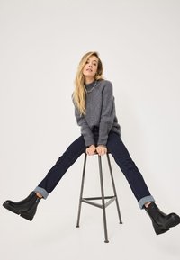 Suéter gris de punto acanalado con un cuello alto, jeans de mezclilla oscuros con puños enrollados y botas negras hasta el tobillo sobre un taburete.