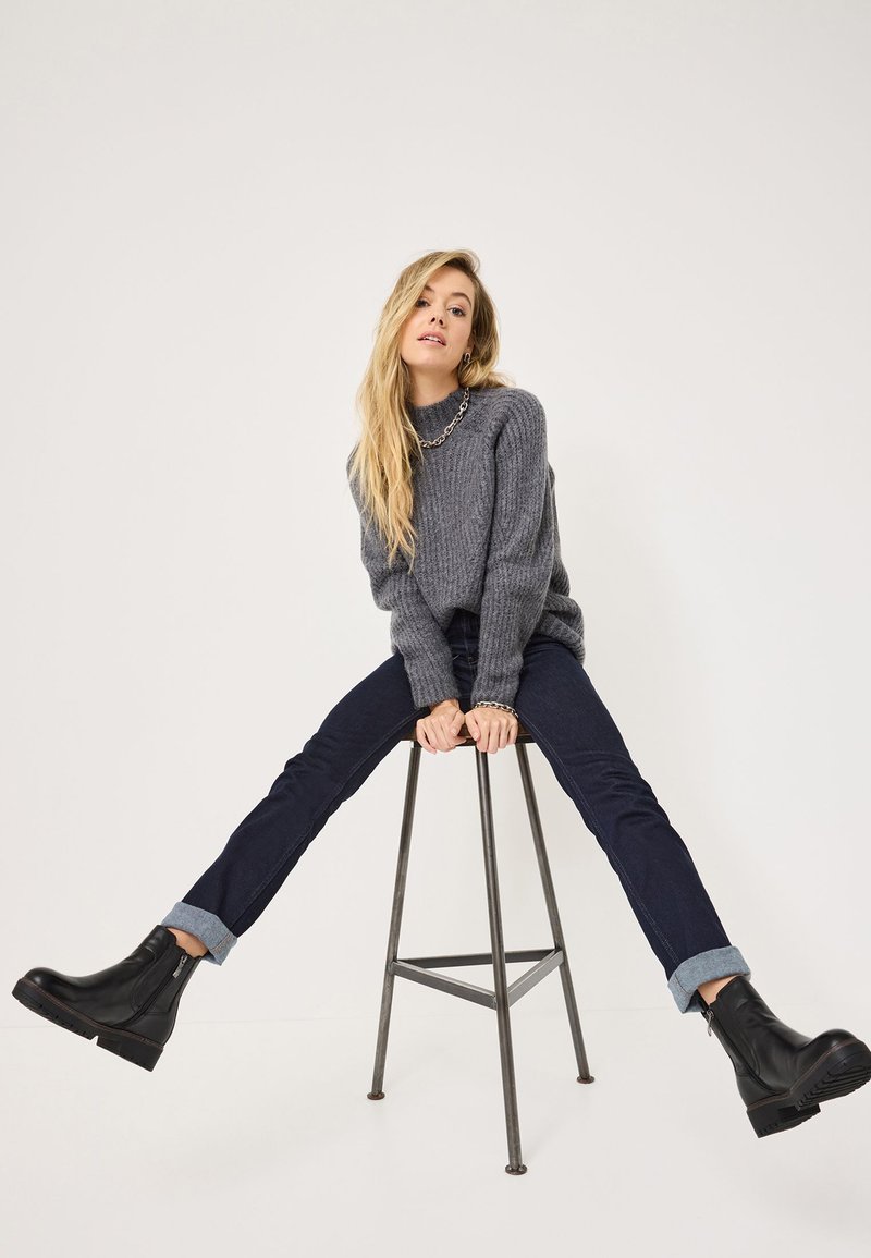 Suéter gris de punto acanalado con un cuello alto, jeans de mezclilla oscuros con puños enrollados y botas negras hasta el tobillo sobre un taburete.