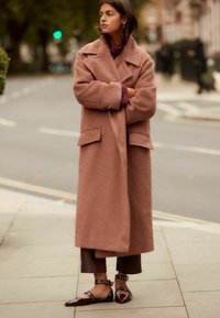 Abrigo oversized de textura, de color marrón claro, con grandes bolsillos, combinado con un suéter burdeos y pantalones marrón oscuro, acompañado de zapatos planos y puntiagudos.
