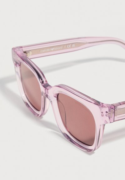 Τετράγωνα γυαλιά ηλίου με διαφανή ροζ σκελετό και φακούς με ροζ απόχρωση που ακουμπούν σε μια λευκή επιφάνεια.