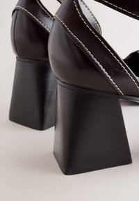 Chaussures à talons blocs en cuir marron foncé avec couture blanche visible et talons larges de forme carrée sur un fond clair.