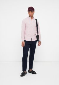 Randstripe rød og hvit skjorte med knapper, marineblå bukser, svarte loafers, og en burgunder cap, med en svart crossbody-veske. Står foran en ensfarget bakgrunn.