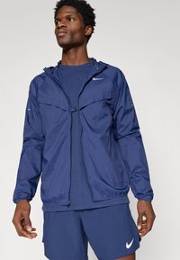 Άνδρας φορώντας μπλε φούτερ με φερμουάρ και κουκούλα, μπλε πουκάμισο και σορτσάκι, με λευκά λογότυπα Nike στο φούτερ και το σορτσάκι.