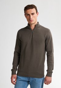 Maglione zip-up di un verde oliva scuro con un motivo testurizzato, polsini e orlo a costine; presenta una zip frontale. Indossato con jeans blu chiaro.