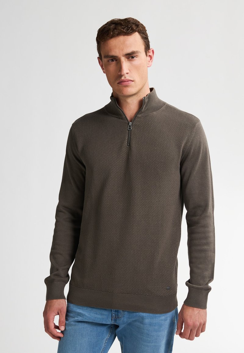 Maglione zip-up di un verde oliva scuro con un motivo testurizzato, polsini e orlo a costine; presenta una zip frontale. Indossato con jeans blu chiaro.