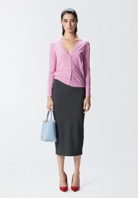 Ružičasti kardigan s v-izrezom i detaljima na gumbima, siva uska olovka suknja, plava torba, i crvene štikle. Glatke teksture i prilagođeni kroj.