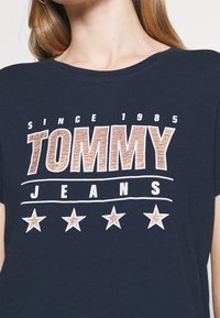 Námořnická modrá bavlněná tričko s texturou loga "TOMMY" v metalicky růžové barvě, s textem "JEANS" a pěti bílými hvězdami pod ním.
