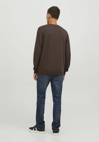 Maglietta marrone a maniche lunghe con polsini e orlo a costine, abbinata a jeans blu scuro. Il modello indossa scarpe da ginnastica nere con suola bianca.