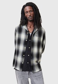Camisa de cuadros en blanco y negro con cuello, que presenta botones dorados y un bolsillo. Lleva puesta sobre una camiseta interior negra, con jeans grises.