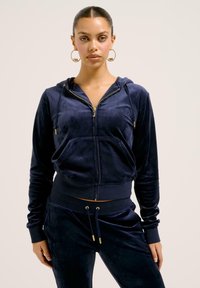 Tuta in velour blu navy con cappuccio corto con zip e cordino, tasche frontali e pantaloni abbinati con dettagli e polsini dorati.