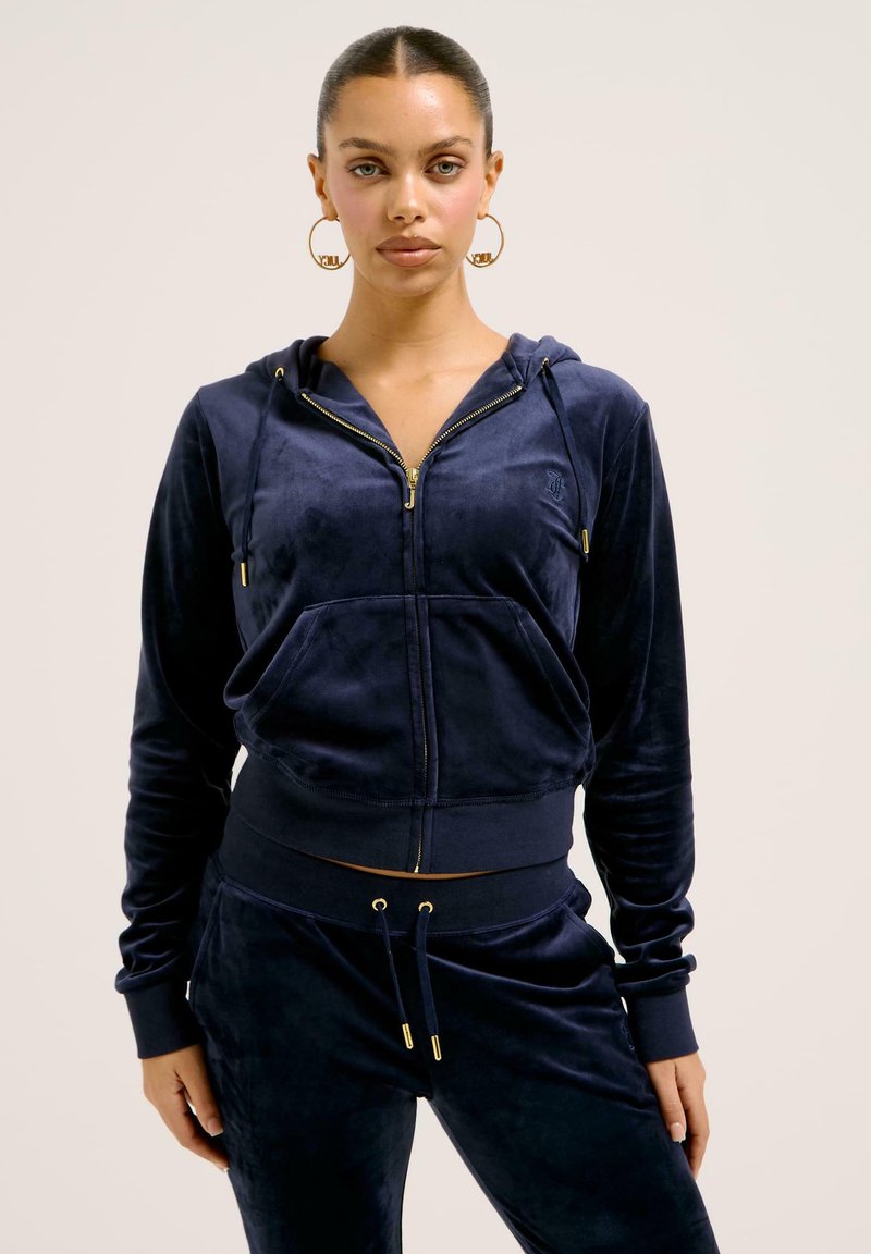 Tuta in velour blu navy con cappuccio corto con zip e cordino, tasche frontali e pantaloni abbinati con dettagli e polsini dorati.