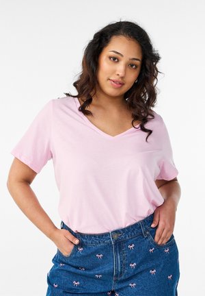 Moteris su bangeliais tamsiais plaukais, vilkinti šviesiai rožinį V formos kaklo marškinėlį ir mėlynas džinsines kelnes, išsiuvinėtas mažais rožiniais kaspinėliais, stovinti su rankomis kišenėse.