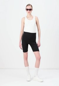 Witte tanktop gecombineerd met hoge zwarte fietsbroek. Model draagt zwarte zonnebril en witte sneakers, wat een slanke silhoutte benadrukt.