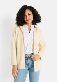Cardigan beige avec capuche, coupe oversize, poignets côtelés et une poche avant, superposé sur une chemise blanche et un jean bleu. Texture en tricot doux.