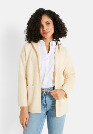 Cárdigan beige con capucha, corte oversized, puños acanalados y un bolsillo frontal, superpuesto sobre una camisa blanca y unos vaqueros azules. Textura de punto suave.