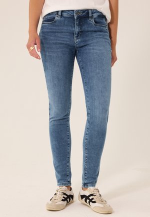 Personne portant un jean skinny bleu clair, une chemise blanche et des baskets blanches à rayures noires, debout devant un fond uni.