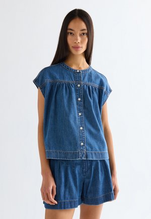 Hosszú egyenes hajjal rendelkező nő, ujjatlan, gombos farmertoppal és hozzáillő farmer rövidnadrággal, sima háttér előtt állva.