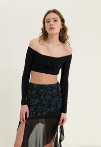 Čierny crop top s odhalenými plecami a detailom uzla, v kombinácii s čiernou sukňou s kvetinovým vzorom, ktorá má priesvitnú vrstvu a rozparok až po stehná.