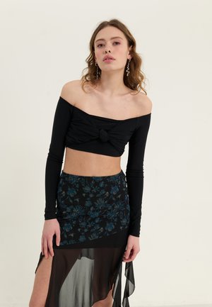 Crni crop top bez naramenica s detaljem čvora, uparen s crnom suknjom s cvjetnim uzorkom koja ima prozirni prekrivač i visoki prorez.