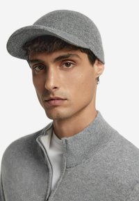 Grå stickad mössa med rund skärm, bärs av en modell. Mjuk textur och åtsittande design, kombinerad med en grå zip-up tröja över en vit skjorta.