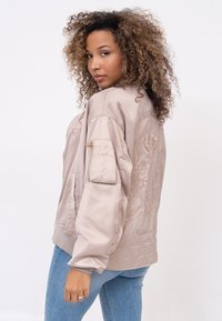 Giacca bomber in morbido satin beige con ricami floreali, polsini elasticizzati e una tasca con cerniera sul braccio. Indossata sopra jeans blu chiaro.