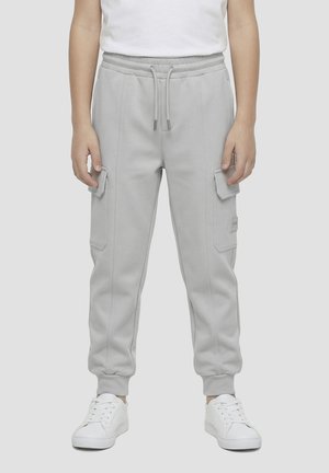 Personne portant un pantalon jogger cargo gris clair avec taille à cordon et poignets élastiques, associé à des baskets blanches et une chemise blanche.