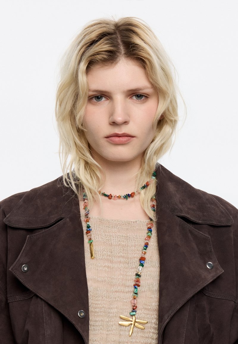 Chocolate brown suede jacket over a beige sweater, featuring a multicolored bead necklace with a gold dragonfly pendant.