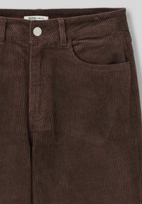 Pantalons larges en velours côtelé marron avec un motif côtelé texturé, comportant une poche avant et une fermeture à bouton métallique au niveau de la taille.