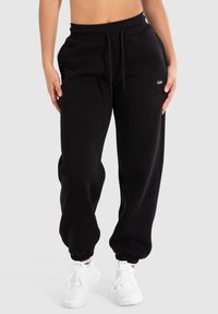 Schwarze Sweatpants mit einem Kordelzugbund, gerippten Knöcheln und einem kleinen gestickten Logo an der linken Hüfte. Getragen mit weißen Sneakern.