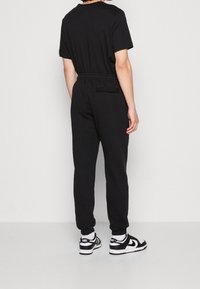 Svart jumpsuit med korta ärmar, elastisk midja och tapered byxor, tillverkad av mjukt tyg. Bärs tillsammans med svart och vita sneakers.