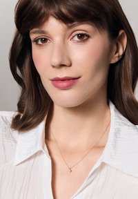 Złoty naszyjnik z małym wisiorkiem, noszony na pomarszczonej białej koszuli z kołnierzykiem, z miękkimi falami i naturalnymi brązowymi włosami.