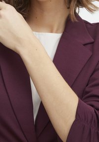 Blazer bordeaux avec des revers structurés et des manches retroussées, porté sur un haut blanc, mettant en valeur un tissu lisse et une coupe ajustée.