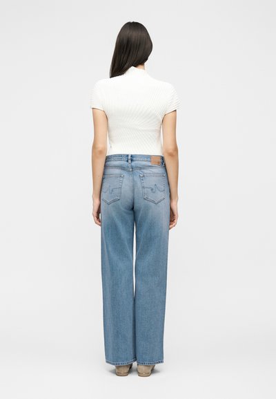 Γυναίκα που στέκεται με την πλάτη στην κάμερα, φοράει λευκό κοντομάνικο μπλουζάκι και ανοιχτό μπλε παντελόνι με φαρδύ πόδι και μπεζ παπούτσια.