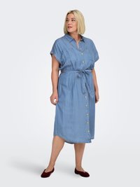 Γυναίκα με φόρεμα midi γαλάζιο, με κουμπιά και δέσιμο στη μέση, με κοντά μανίκια, φοράει σκούρα κόκκινα μπαλαρίνες, στέκεται μπροστά σε απλό φόντο.