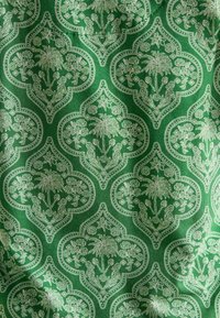 Zaļš audums ar ziedu rakstu, kurā ir palmu un ziedu motīvi, ko ierāmē sarežģīti balti dizaini un punktu akcenti.