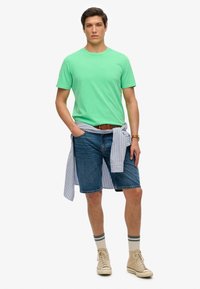 Mintgrünes T-Shirt mit kurzen Ärmeln, blaue Jeansshorts, hellbluse gestreifte Bluse um die Taille gebunden, beige High-Top-Sneakers, cremefarbene Socken mit grauen Streifen.