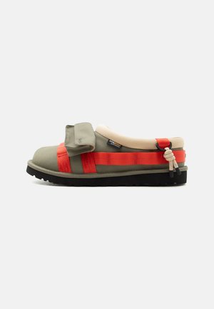 Sandale mule vert olive clair avec lanières rouges, col rembourré beige, semelle noire et détail de rabat avant ajustable sur fond blanc.