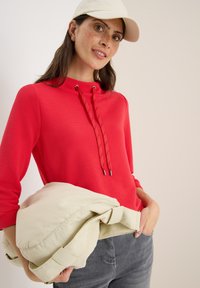 Rotes geripptes Langarmshirt mit hohem Kragen und verstellbaren Kordeln, kombiniert mit einer hellbeigen Jacke, die in einer Hand gehalten wird, und getragen mit grauen Jeans.
