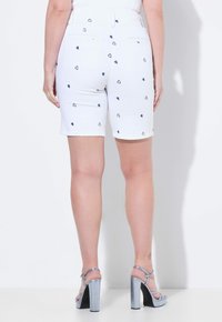 Pantalones cortos de mezclilla blanca con patrones de corazones en navy, cintura media y bolsillos traseros. Combinados con zapatos de tacón bloque con tira en el tobillo en color plateado.